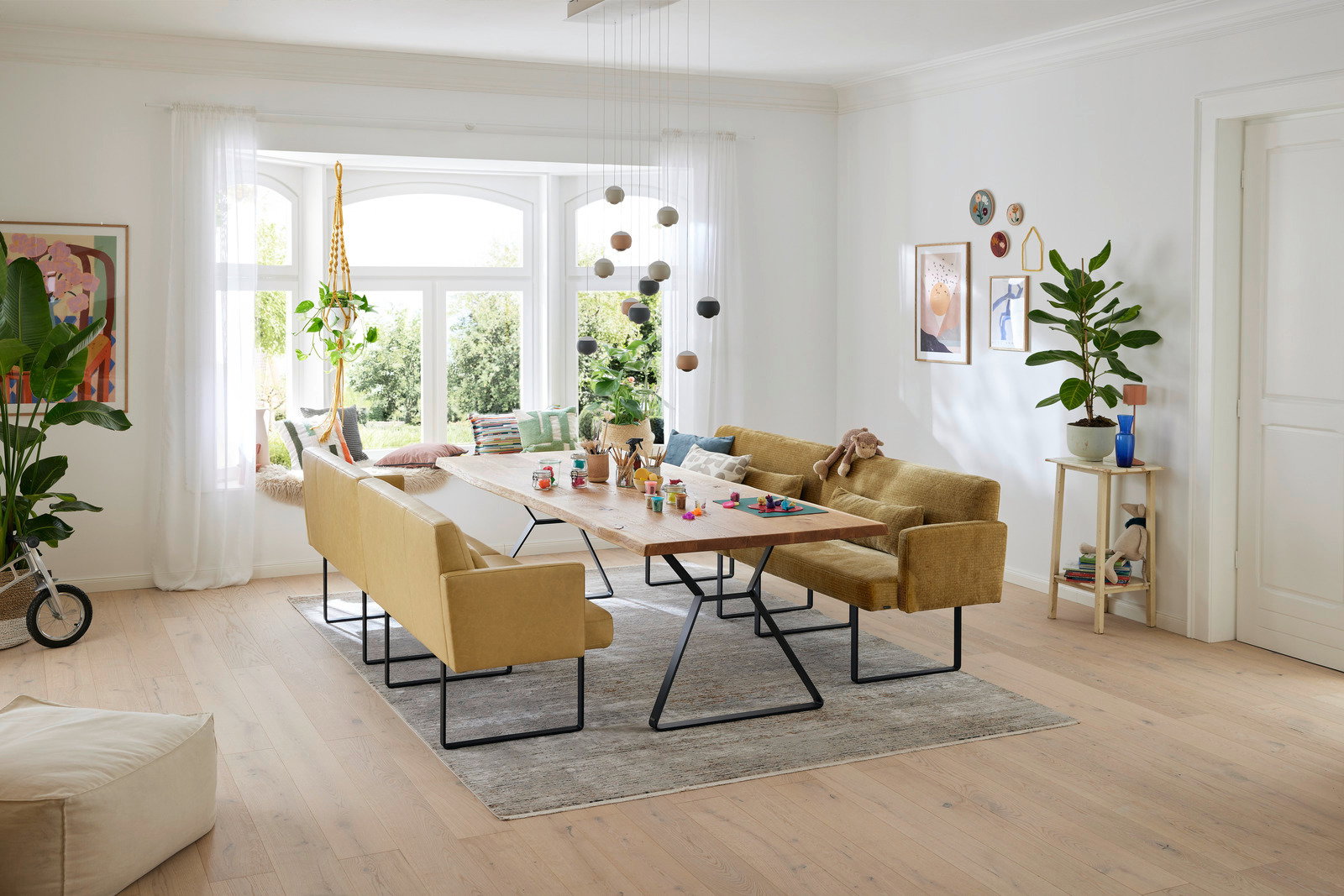 Moderner Esstisch mit Holzplatte und schwarzen Metallbeinen, umgeben von gelben Polsterbänken in einem hellen Wohnzimmer mit großen Fenstern, Blick von der Seite.