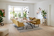 Moderner Esstisch mit Holzplatte und schwarzen Metallbeinen, umgeben von gelben Polsterbänken in einem hellen Wohnzimmer mit großen Fenstern, Blick von der Seite.