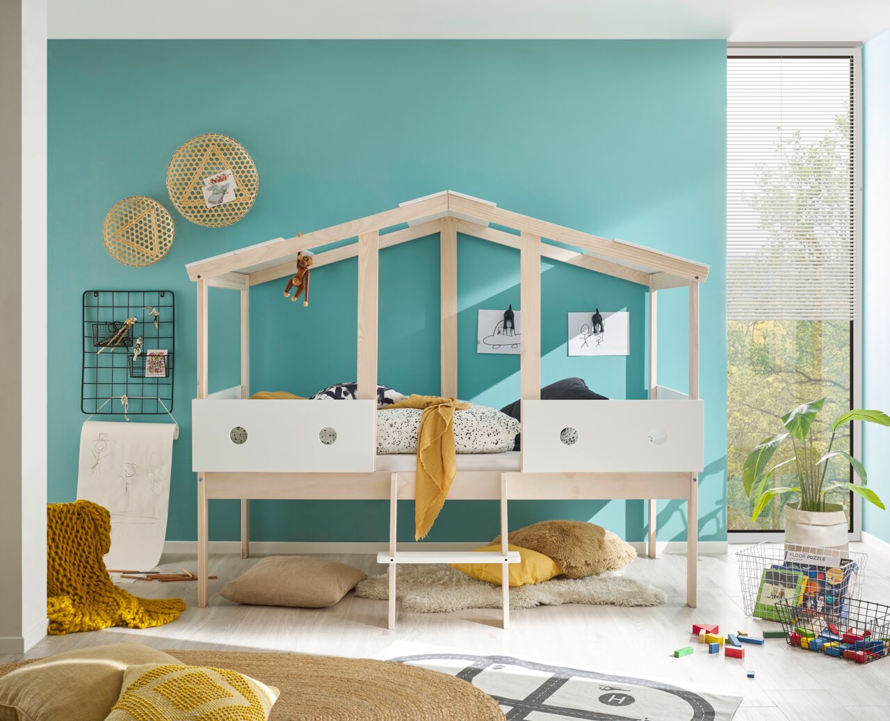 Spielbett in Hausform aus hellem Holz mit weißem Geländer, dekoriert mit Kissen und Decken, in einem Kinderzimmer mit türkisfarbener Wand. Perspektive: Frontalansicht.