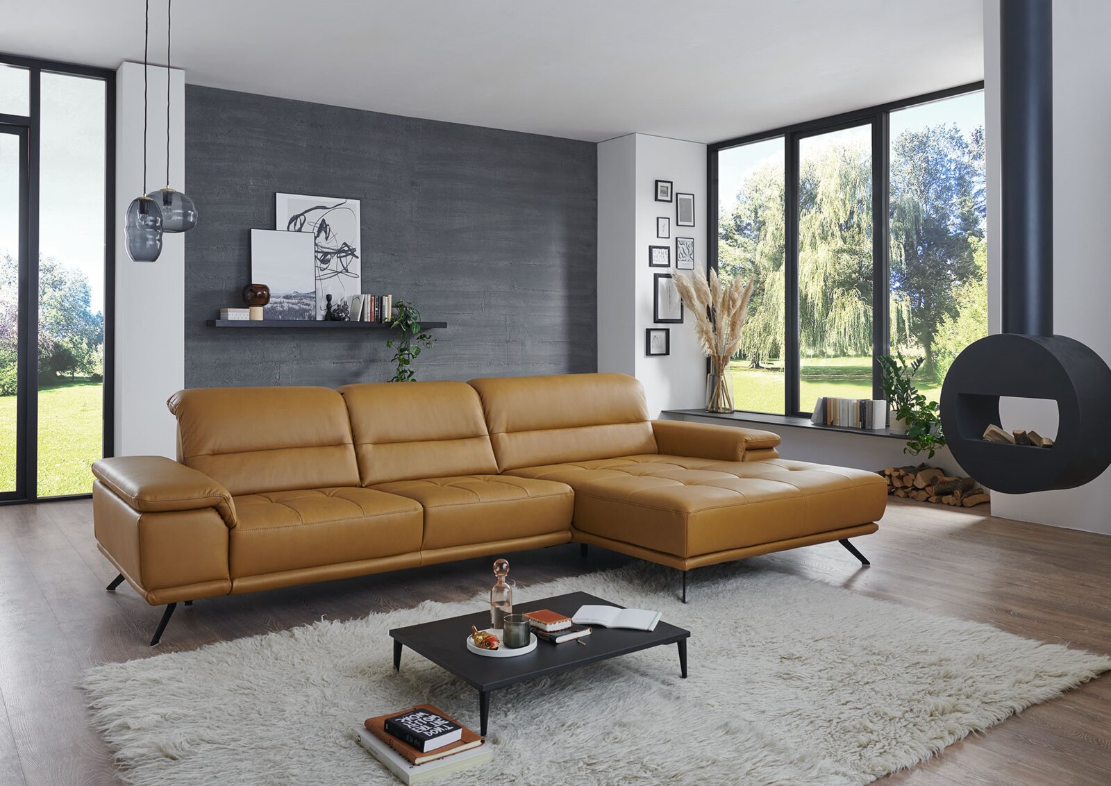 Modernes Wohnzimmer mit cognacfarbenem Leder-Ecksofa mit rechter Ottomane und schwarzen Metallfüßen auf hellem Teppich, aufgenommen aus frontaler leicht schräger Perspektive.