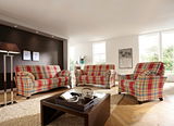 Gemütliches Wohnzimmer mit einem rot-karierten Sessel im Vordergrund, flankiert von einem passenden Sofa und einem weiteren Sessel im Hintergrund. Die Perspektive zeigt den Raum von schräg vorne.