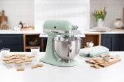 Küchenmaschine Artisan in Pastellgrün auf einer Küchenarbeitsplatte, seitliche Perspektive, umgeben von Keksen und Milchgläsern.