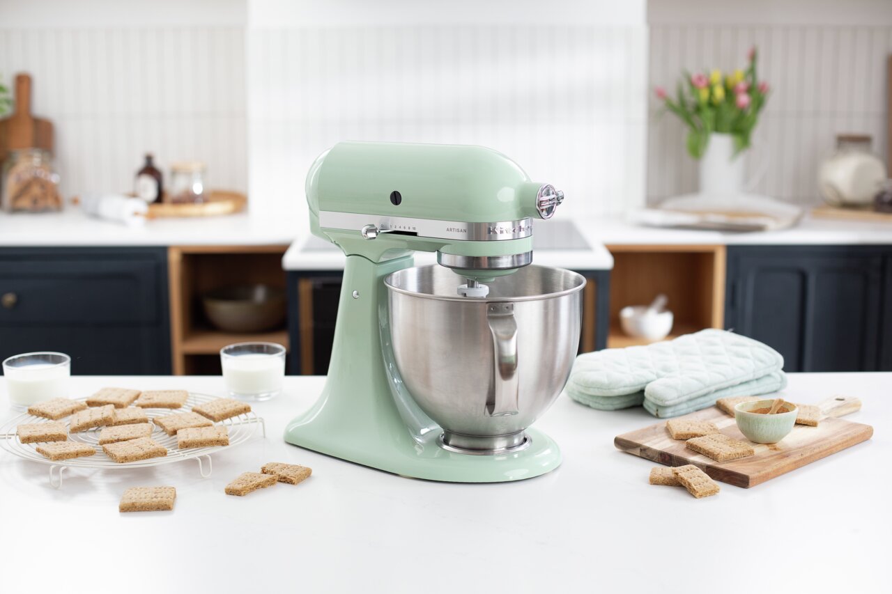 Artisan-keukenmachine in pastelgroen op een keukenblad, gezien vanuit de zijkant, omringd door koekjes en glazen met melk.