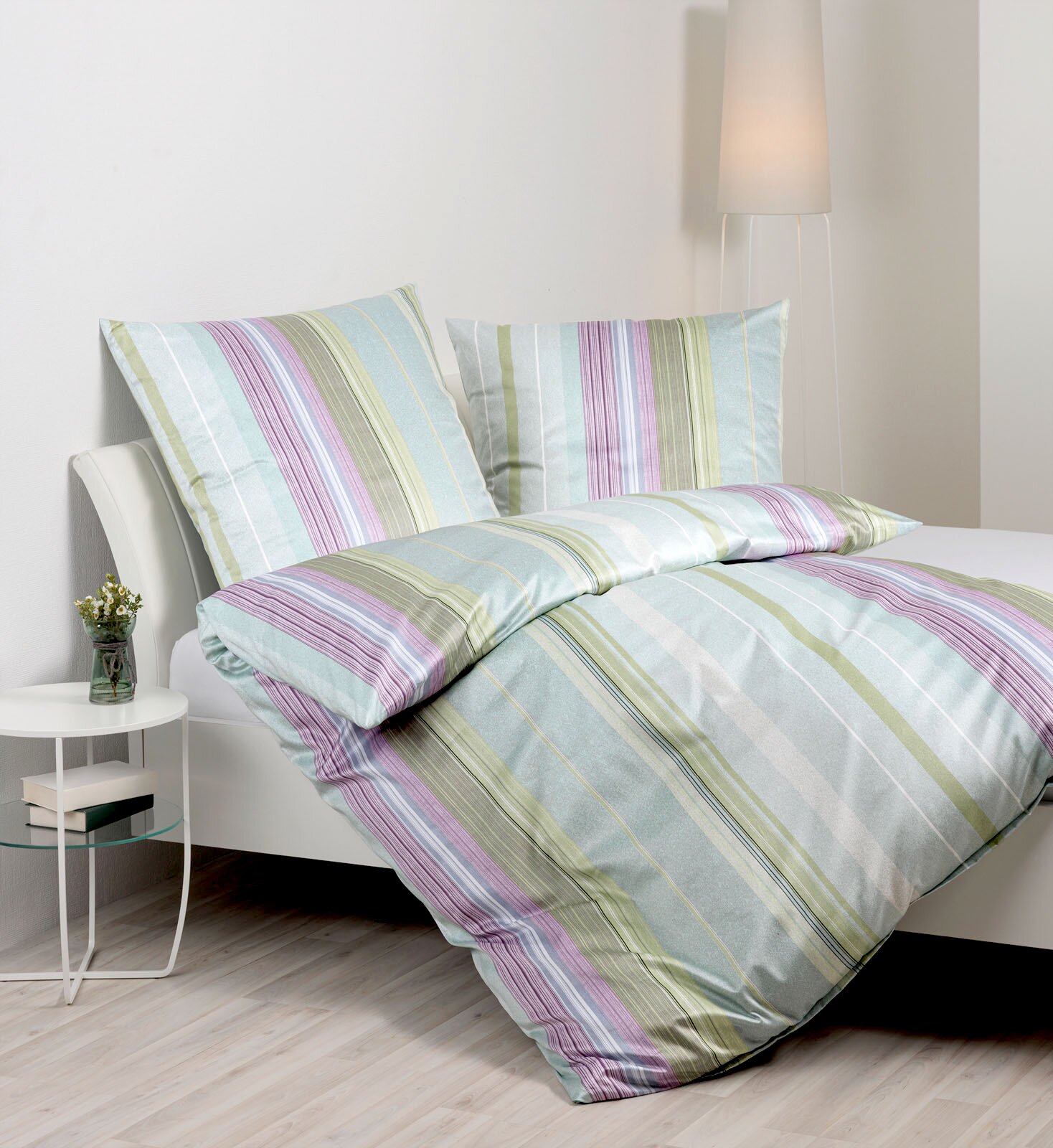 Gestreifte Bettwäsche in Pastellfarben (Mint, Grau, Grün und Lila) mit zwei Kissen und Decke auf einem weißen Bett, fotografiert in leichter Schrägansicht von vorne.