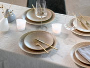 Schlicht gedeckter Esstisch mit beigem, geriffeltem Geschirr mit Goldrand, goldfarbenem Besteck, Weingläsern und Teelichtern auf hellem Leinentischtuch, fotografiert aus leicht erhöhter schräger Perspektive von oben.