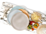 Drei stapelbare Keramikteller in Pastellfarben (blau, orange, creme) auf rustikalem weißem Holztisch, umgeben von Serviette, Löffel, Holzbrett mit Nudeln und bunten Tomaten, Draufsicht (von oben).