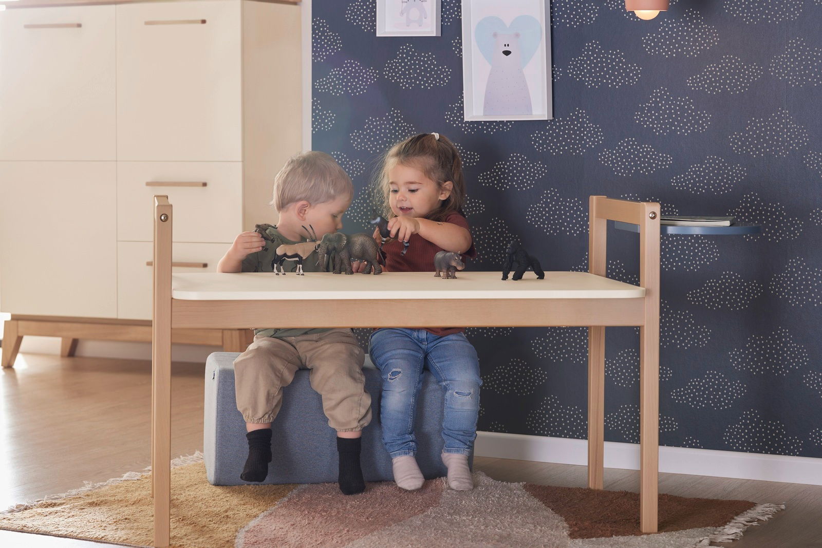 Zwei Kinder spielen mit Tierfiguren auf einer Tischplatte in einem Kinderzimmer. Die Tischplatte ist Teil des Tiny Table, aus hellem Holz, mit einer modernen und minimalistischen Gestaltung. Perspektive: Frontalansicht auf den Tisch.