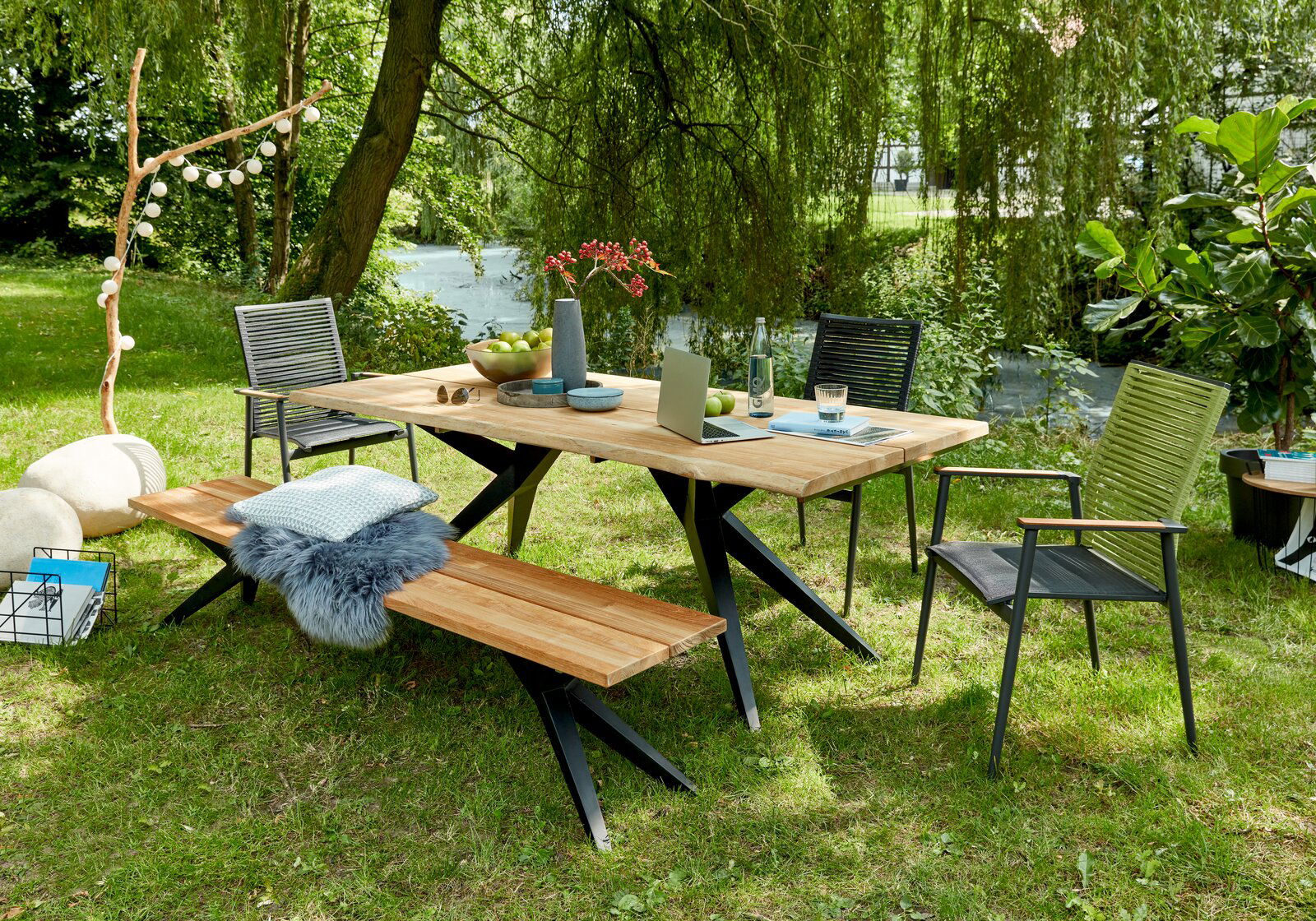 Musterring Esstisch MR HOLLAND Moderner Esstisch im Freien mit Holzplatte und Metallbeinen, umgeben von Stühlen und einer Bank, in einem grünen Garten mit Blick auf einen Fluss.