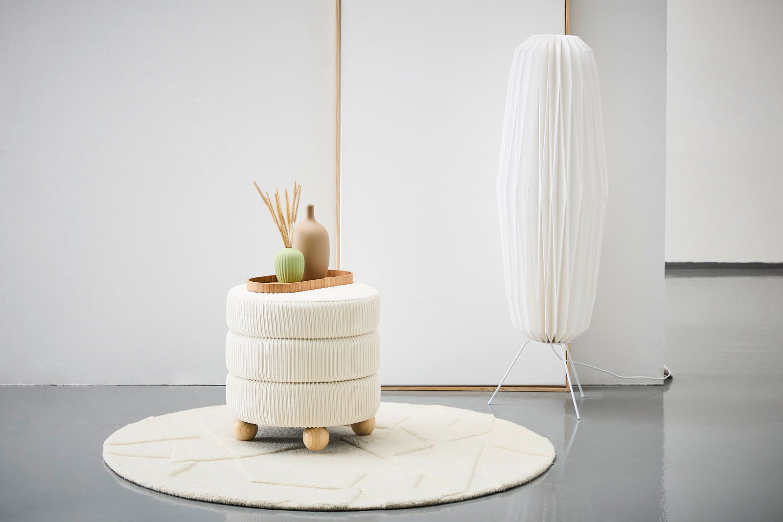 Moderner Hocker mit cremefarbenem Stoffbezug und Holzfüßen, dekoriert mit Vasen auf einem Tablett, auf einem runden Teppich in einem minimalistischen Raum. Seitliche Perspektive.