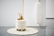 Moderner Hocker mit cremefarbenem Stoffbezug und Holzfüßen, dekoriert mit Vasen auf einem Tablett, auf einem runden Teppich in einem minimalistischen Raum. Seitliche Perspektive.