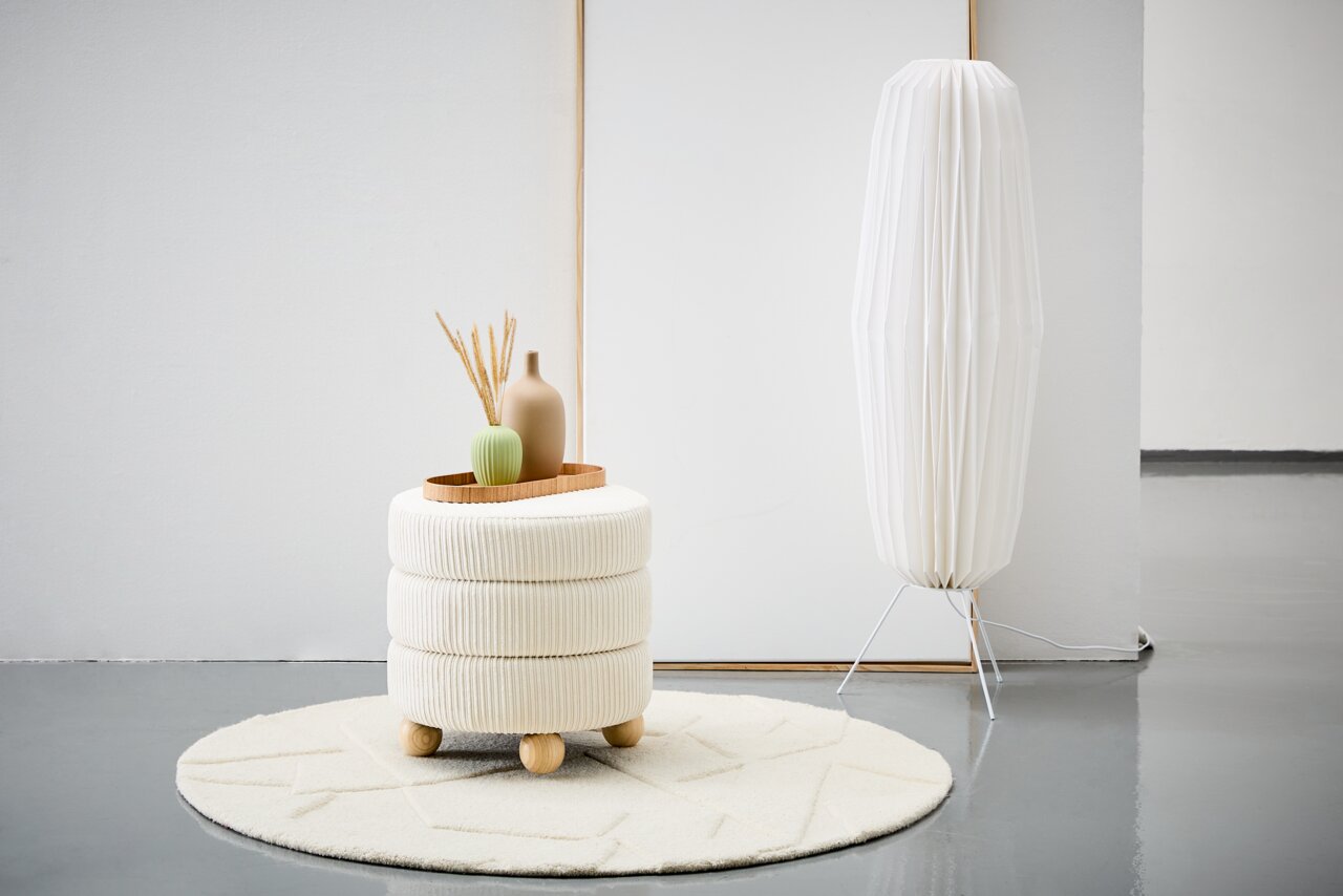 Moderner Hocker mit cremefarbenem Stoffbezug und Holzfüßen, dekoriert mit Vasen auf einem Tablett, auf einem runden Teppich in einem minimalistischen Raum. Seitliche Perspektive.