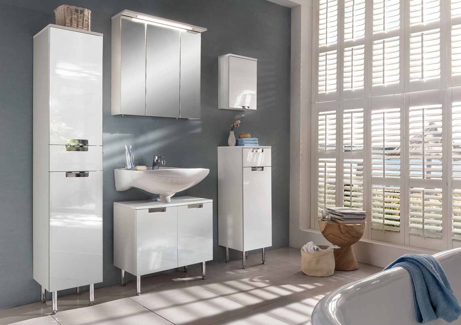 Moderner weißer Hochschrank im Badezimmer, links im Bild, mit glänzender Oberfläche und verchromten Griffen, umgeben von weiteren Badmöbeln und einem großen Fenster mit Jalousien.