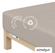 Spannbettlaken XL aus Bio-Baumwolle und Elasthan in Beige, Perspektive von der Ecke des Bettes, mit Hinweis auf 100% Recycling-Verpackung.