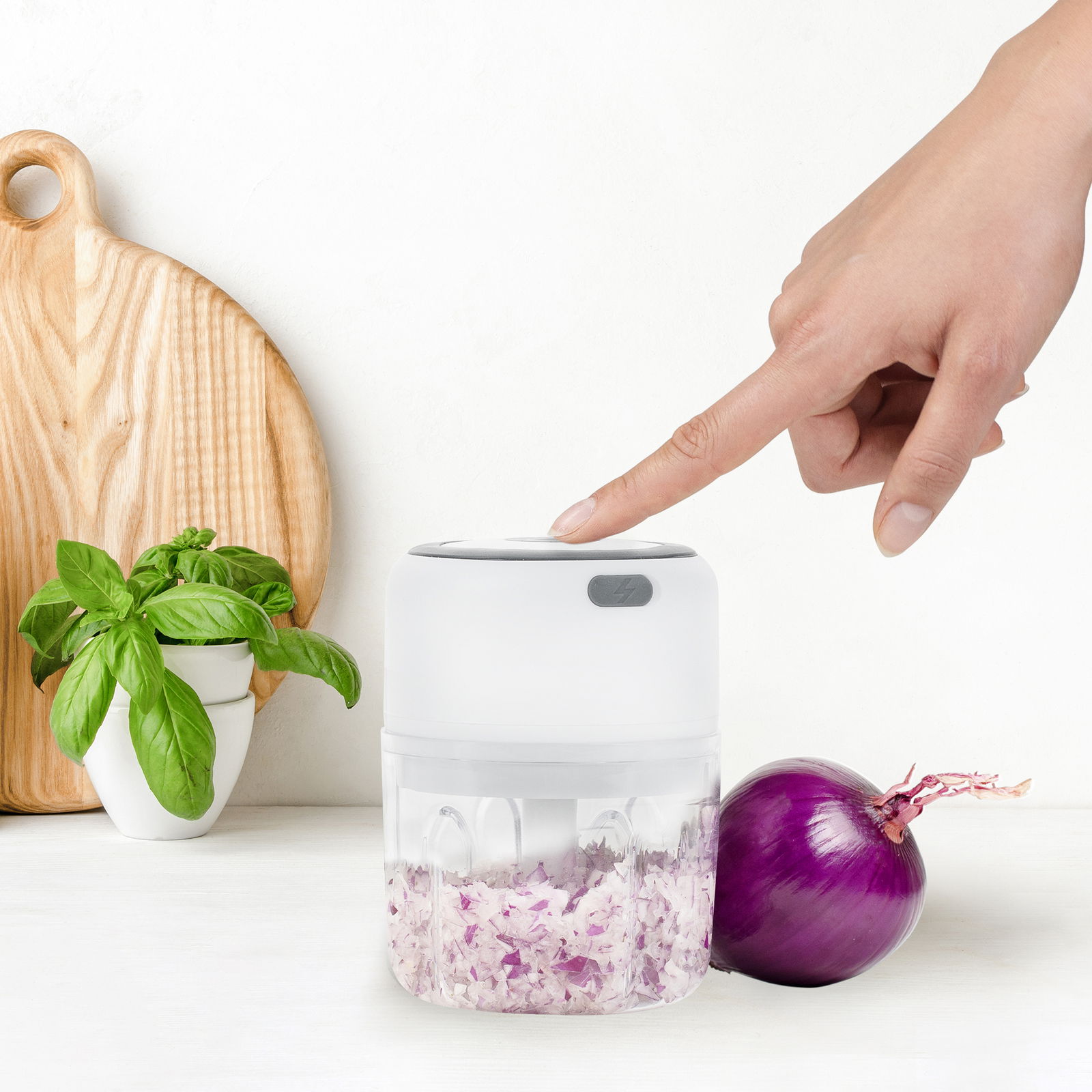 Genius Choptastic 2 Pro NICER DICER Elektrischer Zerkleinerer Choptastic 2 pro, seitliche Ansicht, mit gehackten Zwiebeln im Behälter, Finger drückt auf den Deckel, neben einer roten Zwiebel und einem Basilikumtopf.