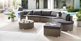 Moderne Garten-Lounge-Möbel in Rattan-Optik mit grauen Kissen, bestehend aus einem Loungesofa, einem Couchtisch mit Glasplatte und einem Hocker, auf einer Terrasse mit Blick auf den Pool.