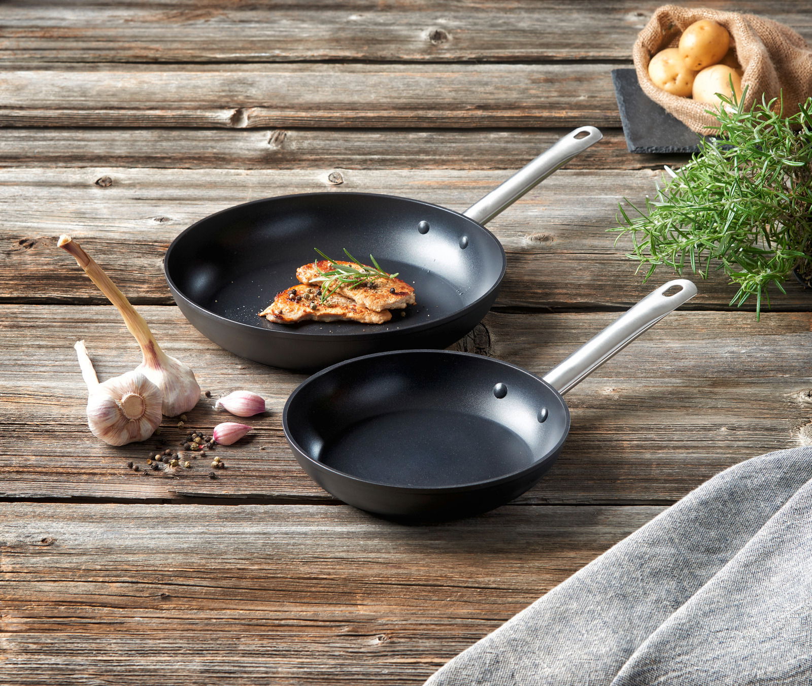 Zwei schwarze Pfannen mit silbernen Griffen auf einem rustikalen Holztisch, eine mit gebratenem Fleisch und Rosmarin, umgeben von Knoblauch, Pfefferkörnern und einem Sack Kartoffeln. Perspektive von oben.