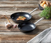 Zwei schwarze Pfannen mit silbernen Griffen auf einem rustikalen Holztisch, eine mit gebratenem Fleisch und Rosmarin, umgeben von Knoblauch, Pfefferkörnern und einem Sack Kartoffeln. Perspektive von oben.