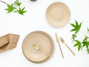 Minimalistisches Geschirr-Arrangement aus zwei beigefarbenen, gerillten Keramiktellern/Schalen mit goldfarbenem Besteck (Löffel, Messer, Gabel) auf weißem Hintergrund, dekoriert mit grünen Blättern und Leinenserviette, Draufsicht (Top-Down).
