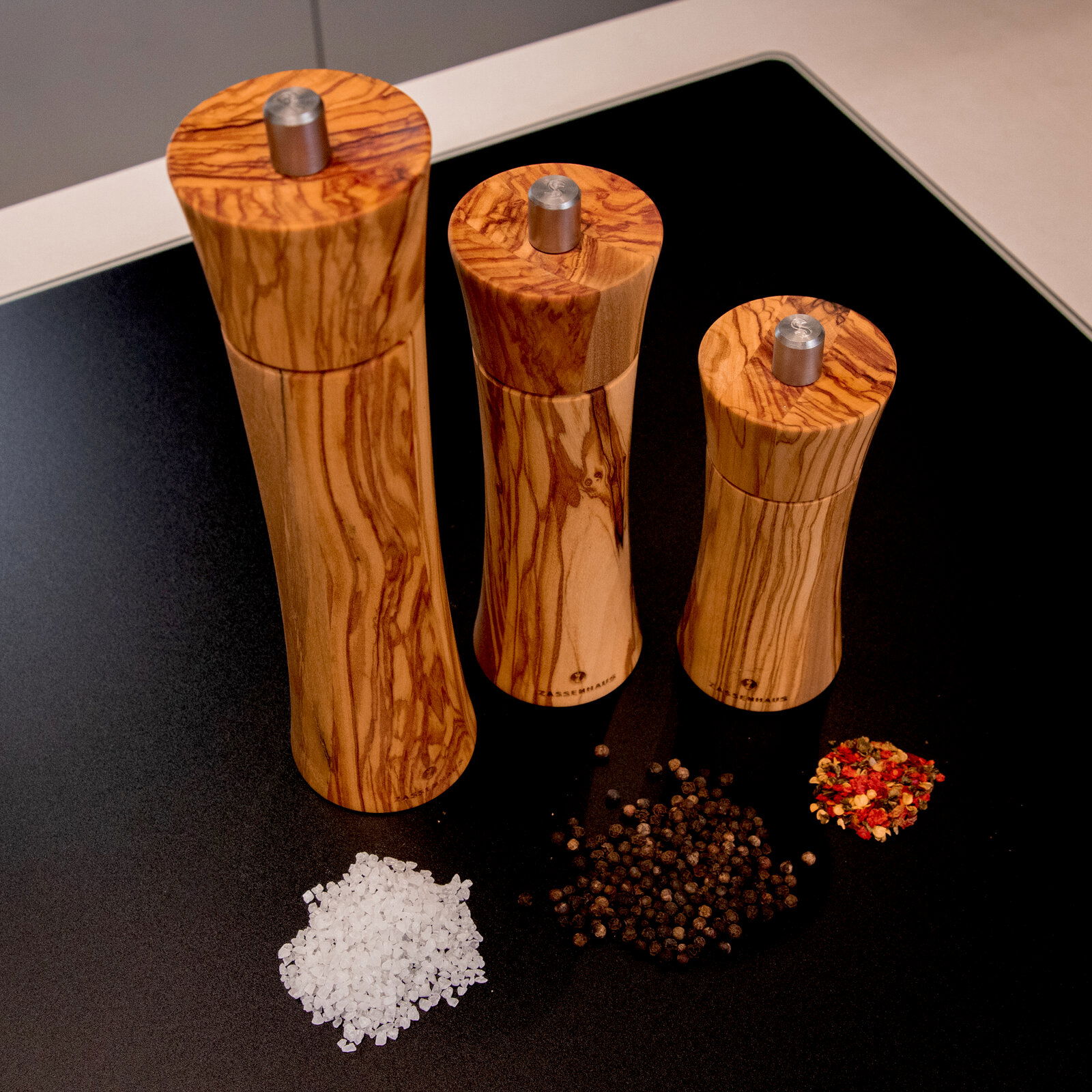 Drei Pfeffermühlen aus Olivenholz in verschiedenen Größen, von oben fotografiert, neben Gewürzen auf einer dunklen Oberfläche.