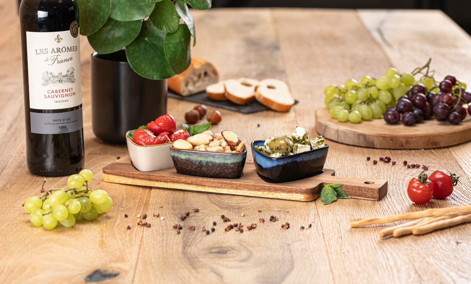 Akazienbrett mit drei Schalen, gefüllt mit Antipasti, auf einem Holztisch, umgeben von Weintrauben, Brot und einer Flasche Rotwein. Perspektive von vorne.