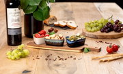 Akazienbrett mit drei Schalen, gefüllt mit Antipasti, auf einem Holztisch, umgeben von Weintrauben, Brot und einer Flasche Rotwein. Perspektive von vorne.