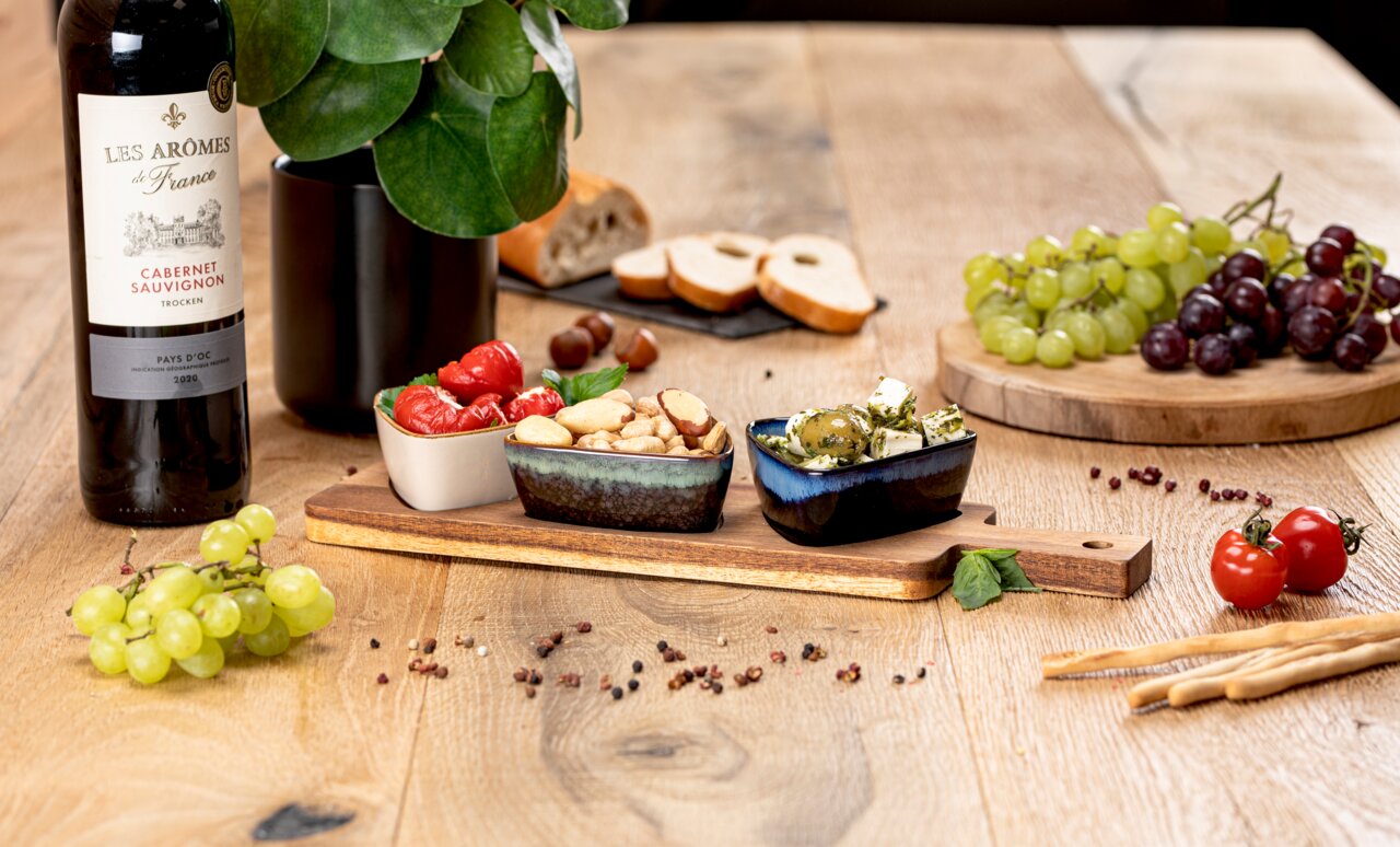 Akazienbrett mit drei Schalen, gefüllt mit Antipasti, auf einem Holztisch, umgeben von Weintrauben, Brot und einer Flasche Rotwein. Perspektive von vorne.