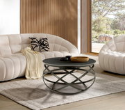 Moderner Couchtisch mit schwarzer runder Tischplatte und kunstvollem Metallgestell, frontal im Wohnzimmer mit weißen Sofas und Holzwand.