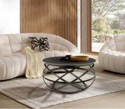Moderner Couchtisch mit schwarzer runder Tischplatte und kunstvollem Metallgestell, frontal im Wohnzimmer mit weißen Sofas und Holzwand.