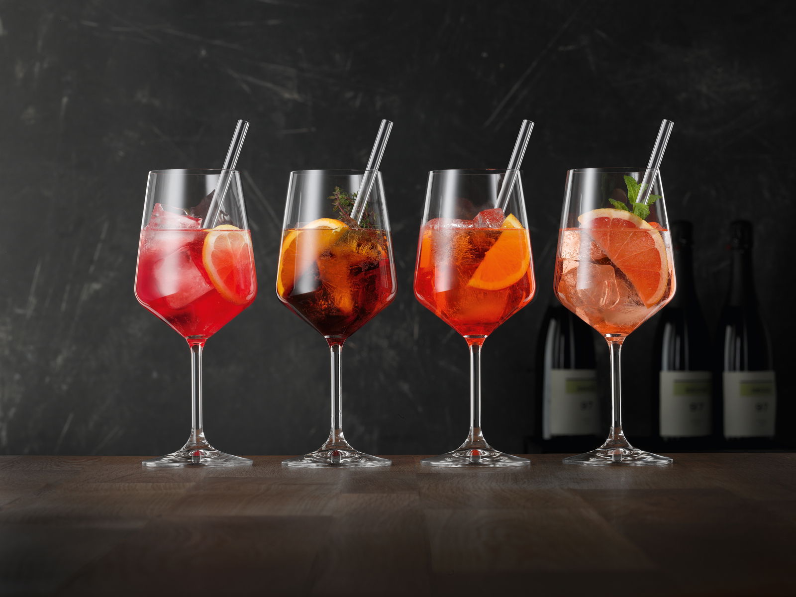 Vier elegante Weingläser mit bunten Cocktails und Eiswürfeln, dekoriert mit Zitrusfrüchten und Kräutern, auf einem Holztisch vor dunklem Hintergrund. Seitliche Perspektive.