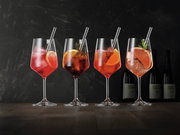 Vier elegante Weingläser mit bunten Cocktails und Eiswürfeln, dekoriert mit Zitrusfrüchten und Kräutern, auf einem Holztisch vor dunklem Hintergrund. Seitliche Perspektive.