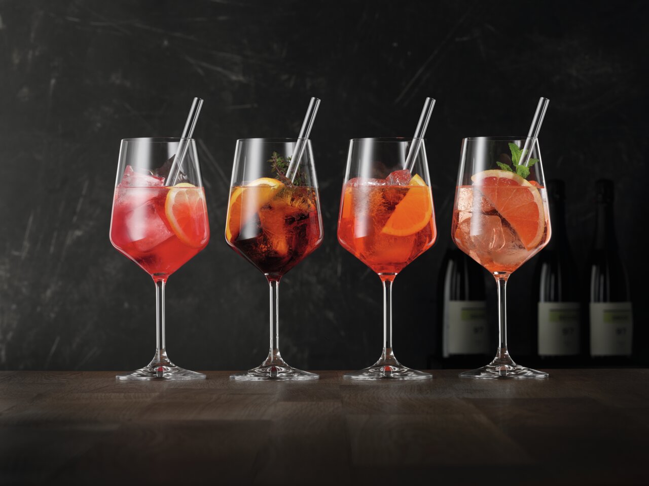 Vier elegante Weingläser mit bunten Cocktails und Eiswürfeln, dekoriert mit Zitrusfrüchten und Kräutern, auf einem Holztisch vor dunklem Hintergrund. Seitliche Perspektive.