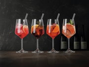 Vier elegante Weingläser mit bunten Cocktails und Eiswürfeln, dekoriert mit Zitrusfrüchten und Kräutern, auf einem Holztisch vor dunklem Hintergrund. Seitliche Perspektive.