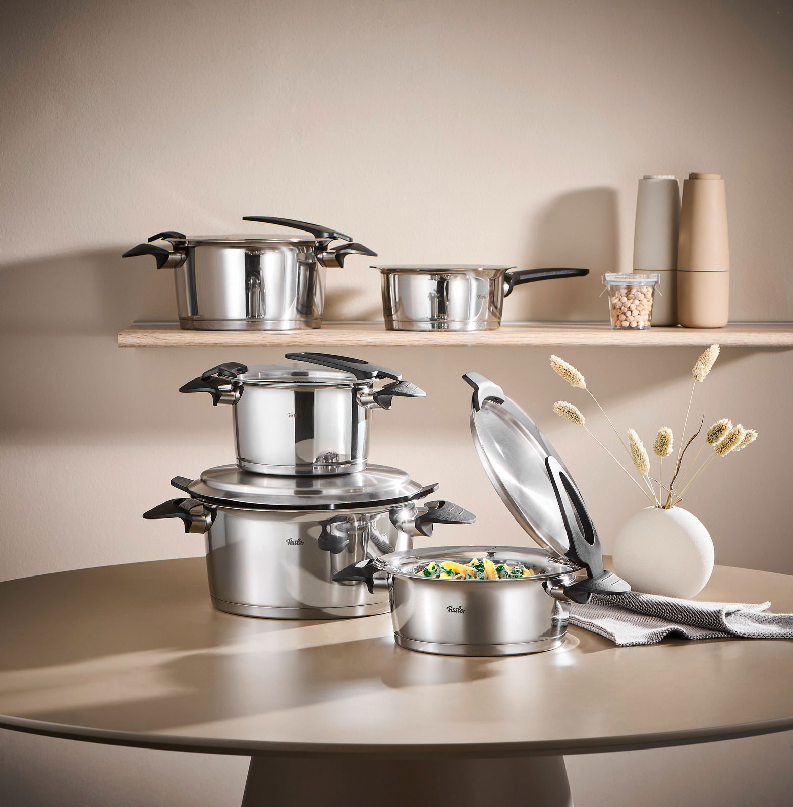 Topfset Intensa 5-tlg. aus Edelstahl, bestehend aus mehreren Töpfen mit Deckeln, auf einem Tisch und einem Regal präsentiert. Perspektive: seitlich.