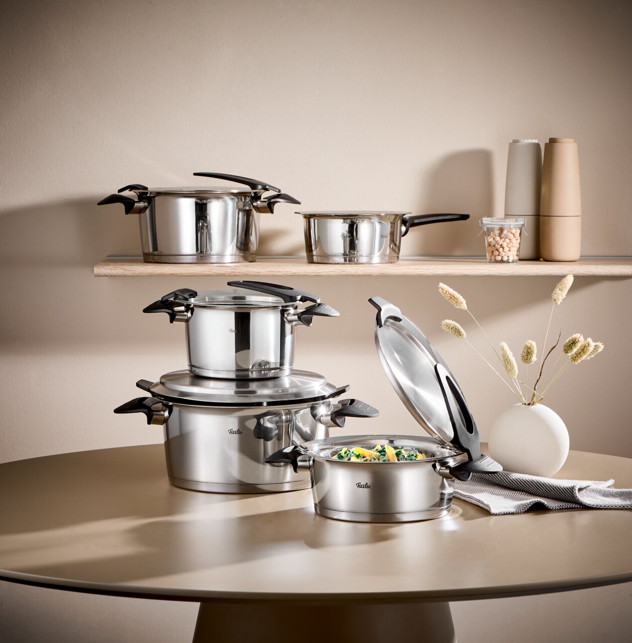 Topfset Intensa 5-tlg. aus Edelstahl, bestehend aus mehreren Töpfen mit Deckeln, auf einem Tisch und einem Regal präsentiert. Perspektive: seitlich.