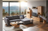Gemütliche Wohnzimmer-Ecke mit grauem Ecksofa und Holzmöbeln, Blick auf Berge und See durch große Fenster.