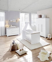 Helles und modernes Babyzimmer mit Kinderbett, Wickelkommode und Kleiderschrank in weißer Farbe, aus der Perspektive eines diagonalen Blicks auf den Raum.