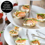 Dreistufige weiße Etagere aus hochwertigem Porzellan, von oben fotografiert, mit belegten Häppchen und Erdbeeren im Hintergrund.