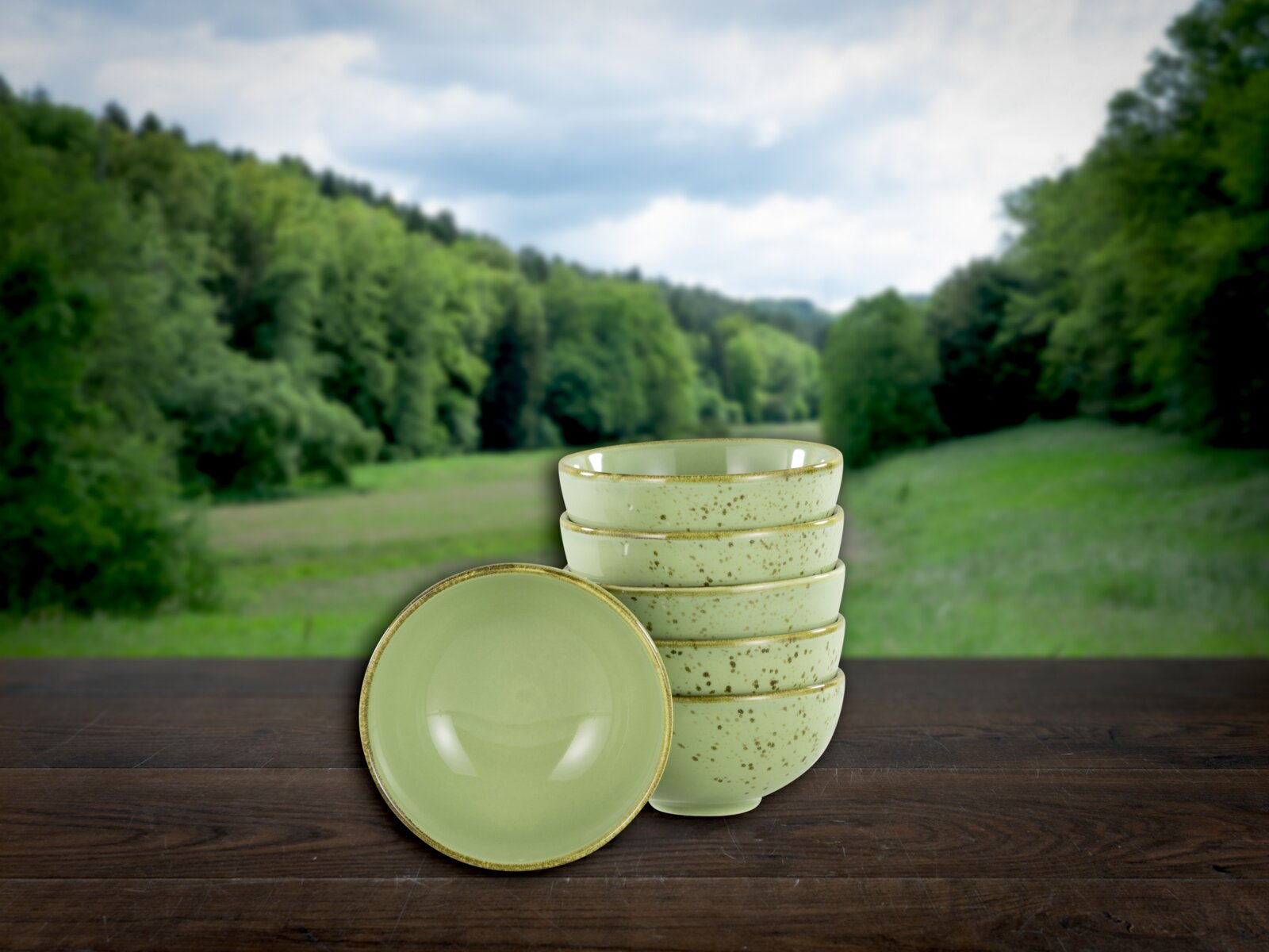 Stapel aus fünf gesprenkelten, hellgrünen Keramikschalen mit einer Schale vorne liegend, auf Holztisch vor unscharfer Wald- und Wiesenlandschaft, Frontansicht leicht von oben