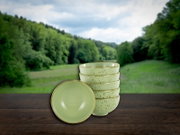 Stapel aus fünf gesprenkelten, hellgrünen Keramikschalen mit einer Schale vorne liegend, auf Holztisch vor unscharfer Wald- und Wiesenlandschaft, Frontansicht leicht von oben