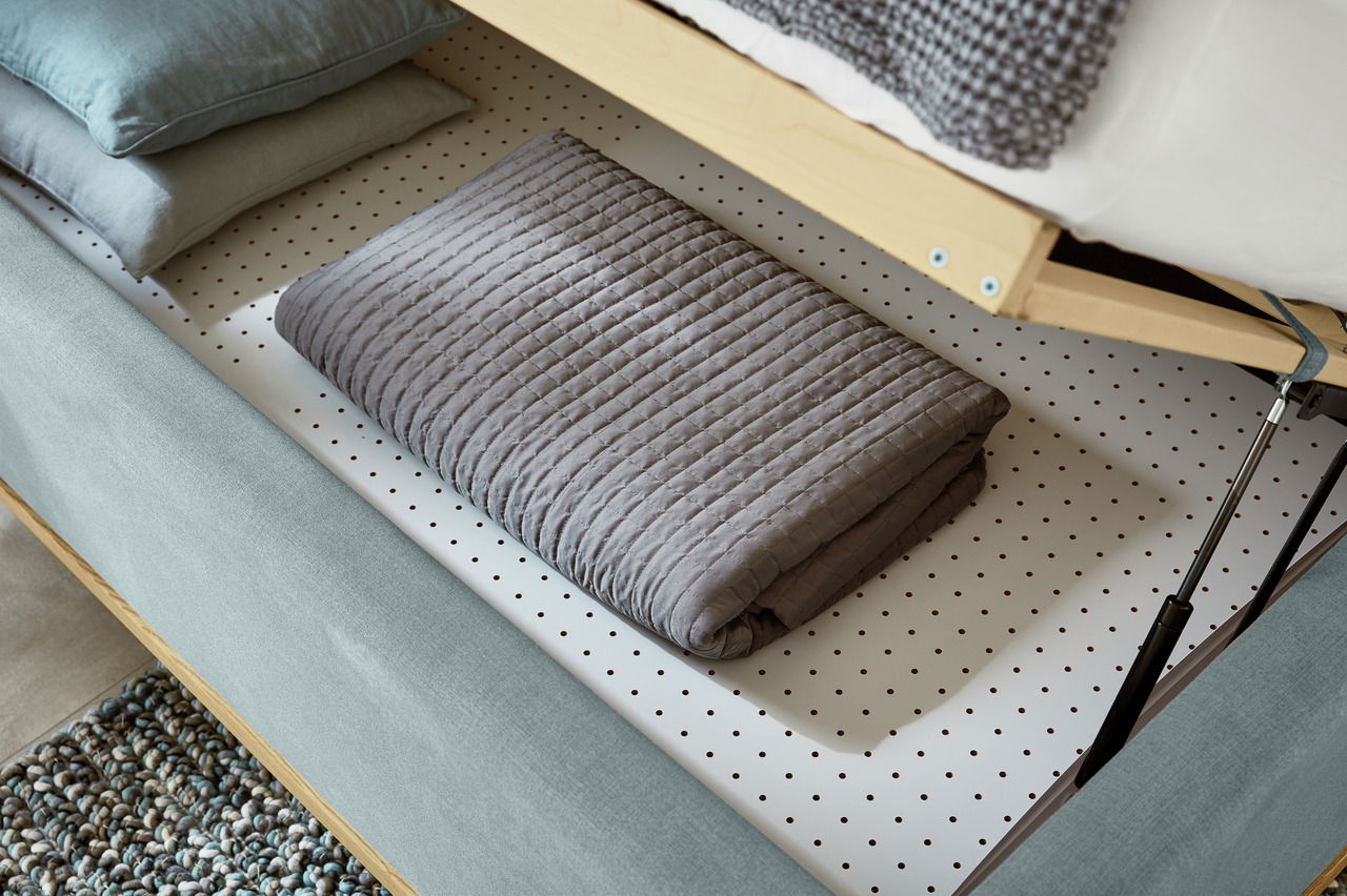 Detailansicht eines Polsterbetts mit geöffnetem Bettkasten, der eine gefaltete graue Decke und Kissen zeigt.