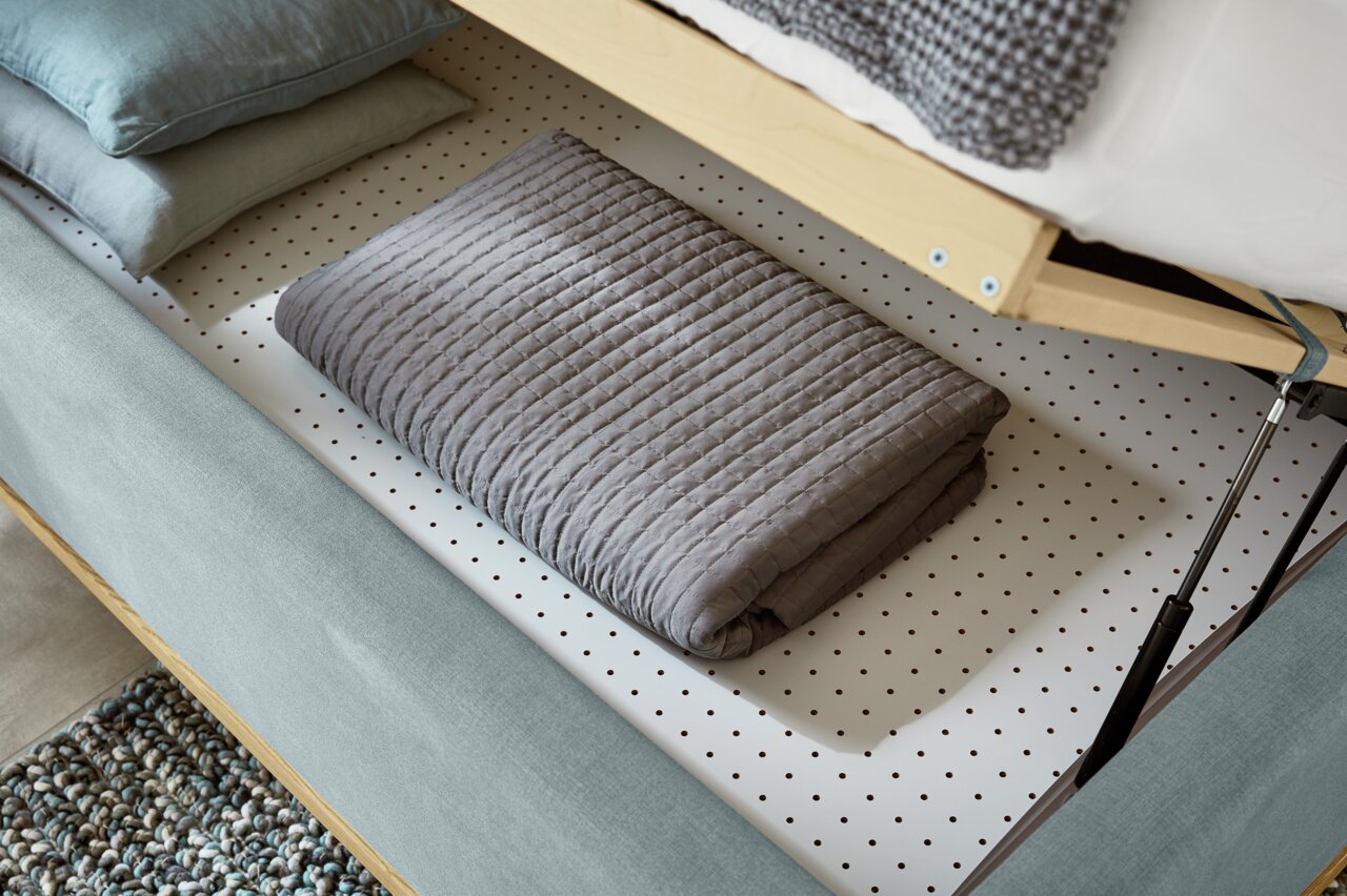 Detailfoto van een gestoffeerd bed met geopende opbergruimte, waarin een opgevouwen grijze deken en kussens te zien zijn.