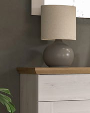 Tischlampe mit rundem, grauem Sockel und beigem Lampenschirm auf einer Holzkommode, seitliche Perspektive