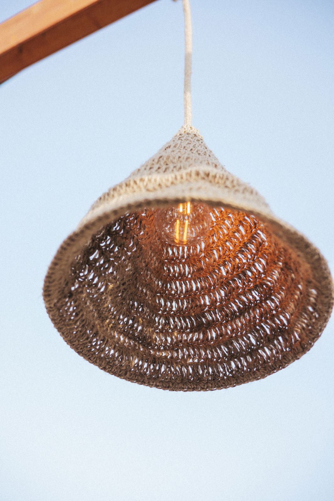 Outdoor-Hängeleuchte aus geflochtenem Material, von unten fotografiert, mit sichtbarer Glühbirne und blauem Himmel im Hintergrund.