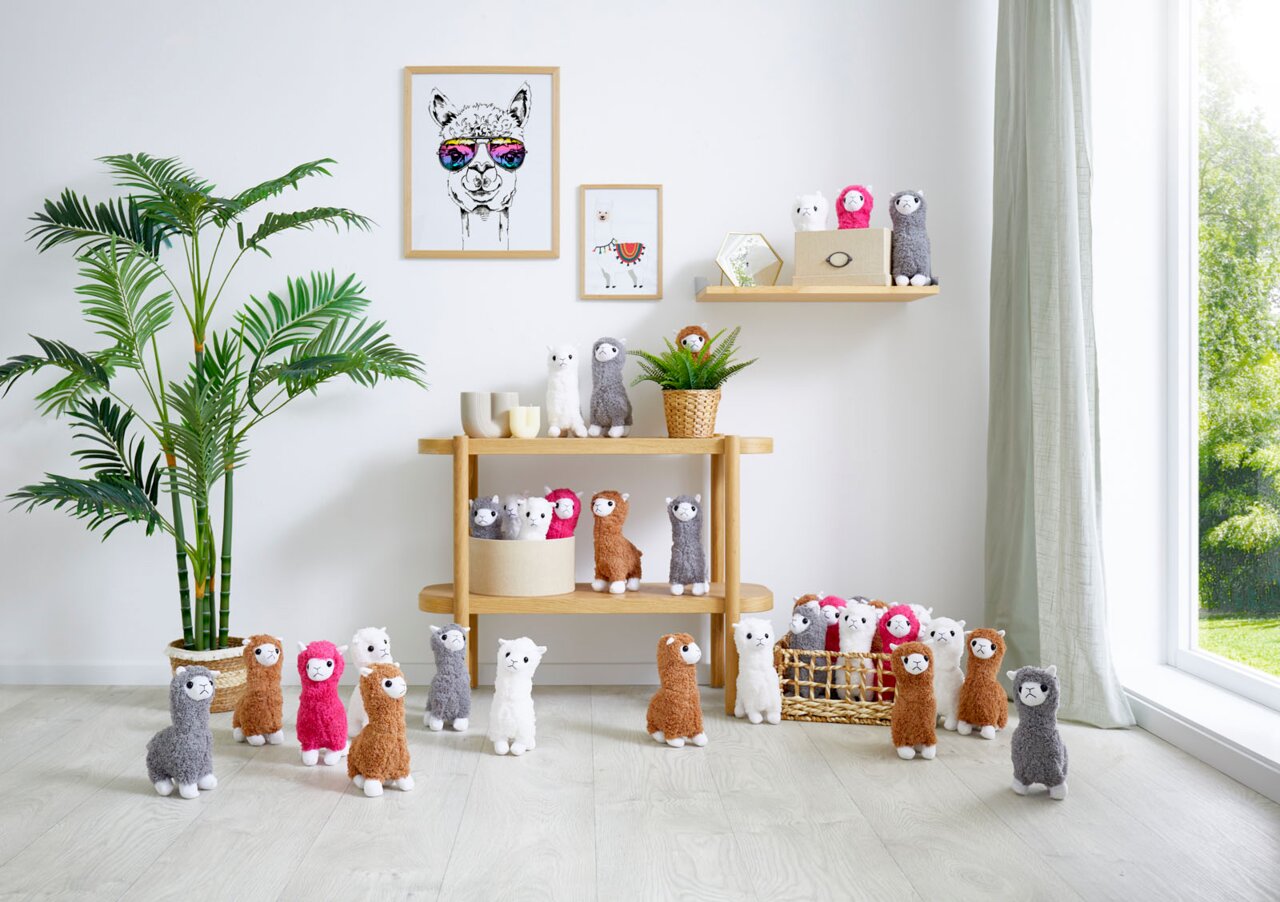 Verschiedene Stofftiere in Form von Alpakas in einem hellen Raum, auf Regalen und dem Boden arrangiert. Perspektive von vorne.