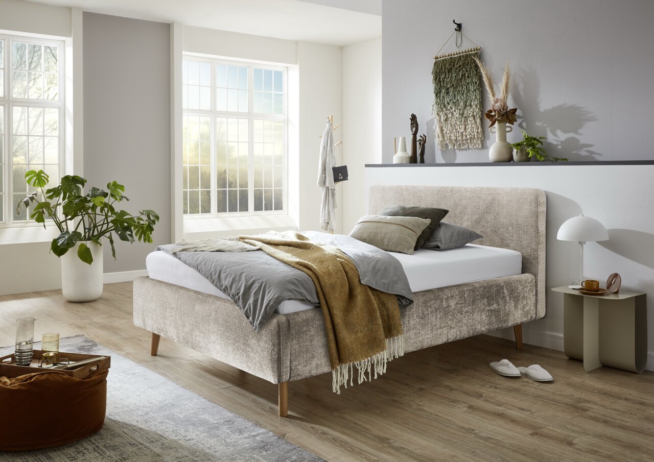 Modernes Polsterbettgestell in einem hellen Schlafzimmer mit großen Fenstern, seitliche Perspektive.