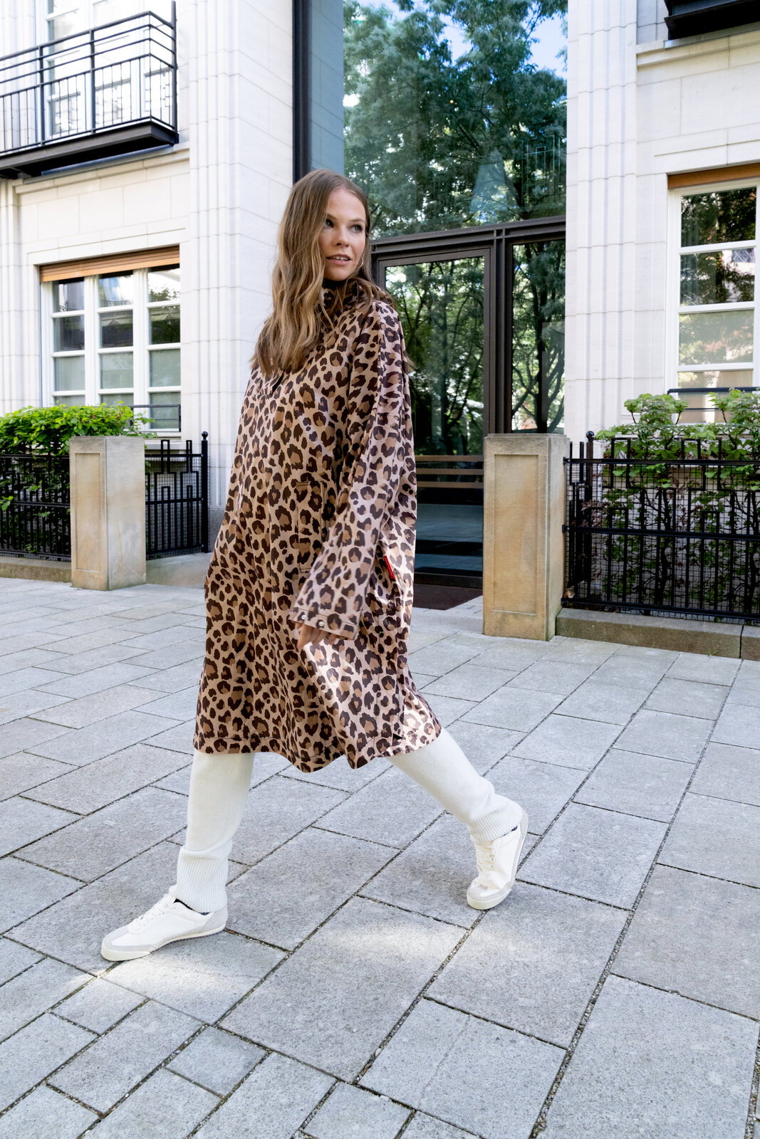 Frau trägt ein knielanges, locker fallendes Kleid mit Leopardenmuster und langen Ärmeln, kombiniert mit weißen Leggings und weißen Sneakern, aufgenommen in einer Ganzkörper-Frontansicht leicht seitlich auf einem Gehweg vor einem modernen Gebäude.