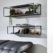 Zwei schwarze, rechteckige Wandregale aus Metall mit Deko (Pflanzen, Bücher) an weißer Wand, schräg von vorn in leichter Seitenperspektive fotografiert; unten ist ein graues Ledersofa angeschnitten sichtbar.