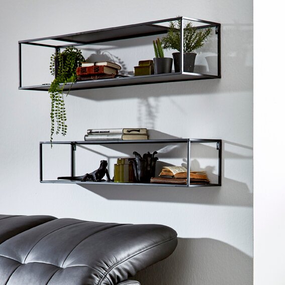 Zwei schwarze, rechteckige Wandregale aus Metall mit Deko (Pflanzen, Bücher) an weißer Wand, schräg von vorn in leichter Seitenperspektive fotografiert unten ist ein graues Ledersofa angeschnitten sichtbar.