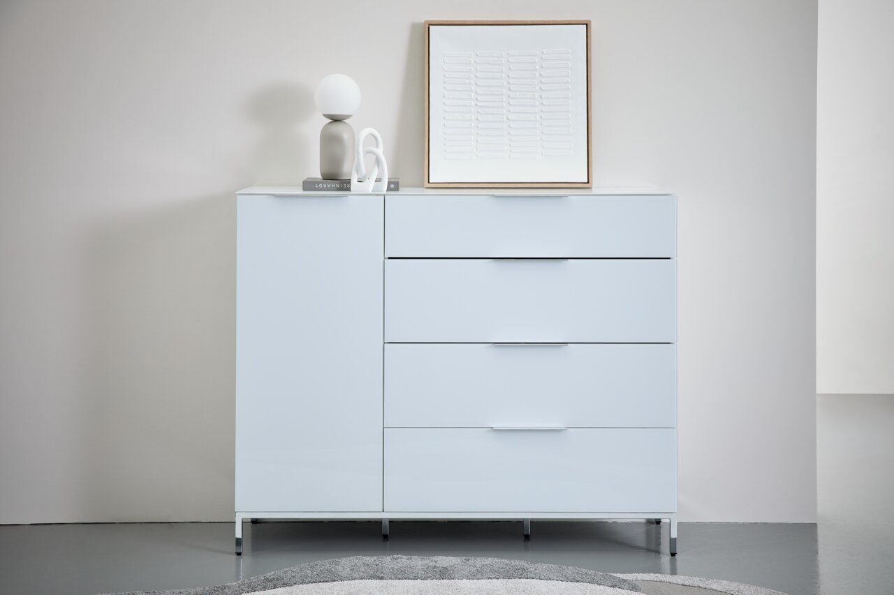 Moderne weiße Kombikommode mit vier Schubladen und einer Tür, frontal fotografiert. Dekoriert mit einer Lampe, einem Buch und einem Kunstwerk auf der Oberseite.