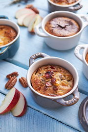 Vogelperspektive auf ein Set von Mini-Cocottes in verschiedenen Farben, gefüllt mit gebackenem Dessert, umgeben von Apfelscheiben und Pekannüssen auf einem blauen Holztisch.